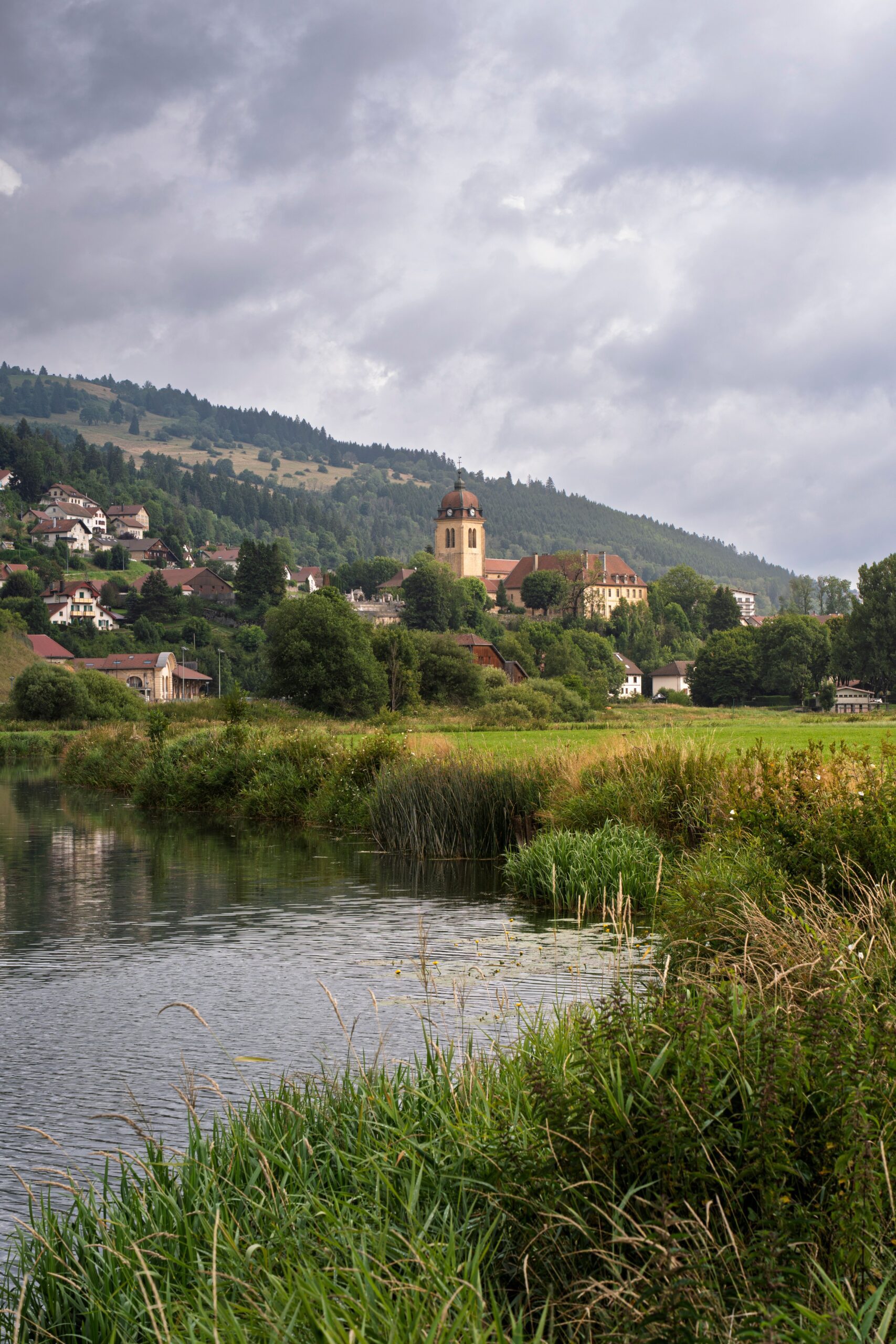Village du Haut-Doubs avec clocher d'église au bord d'une rivière sous un ciel nuageux